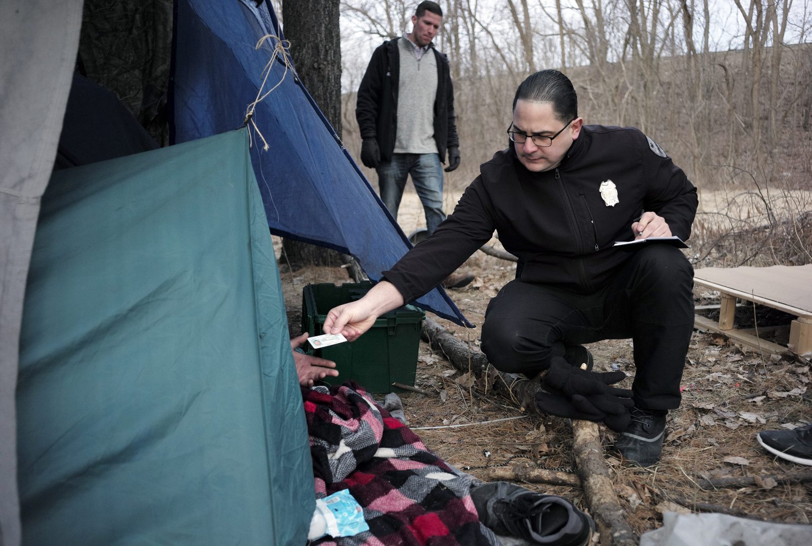 In this Tuesday, Feb. 12, 2019 photo Worcester Police officer Angel Rivera, right, returns a license to an unidentified man as Rivera asks if he has been tested for Hepatitis A at the entrance to a tent where the man spent the night in a wooded area, in Worcester, Mass. Dan Cahill, City of Worcester sanitary inspector, walks behind center. The city was hit hard when recent hepatitis A outbreaks across the country started sickening and killing homeless people and illicit drug users. (AP Photo/Steven Senne)