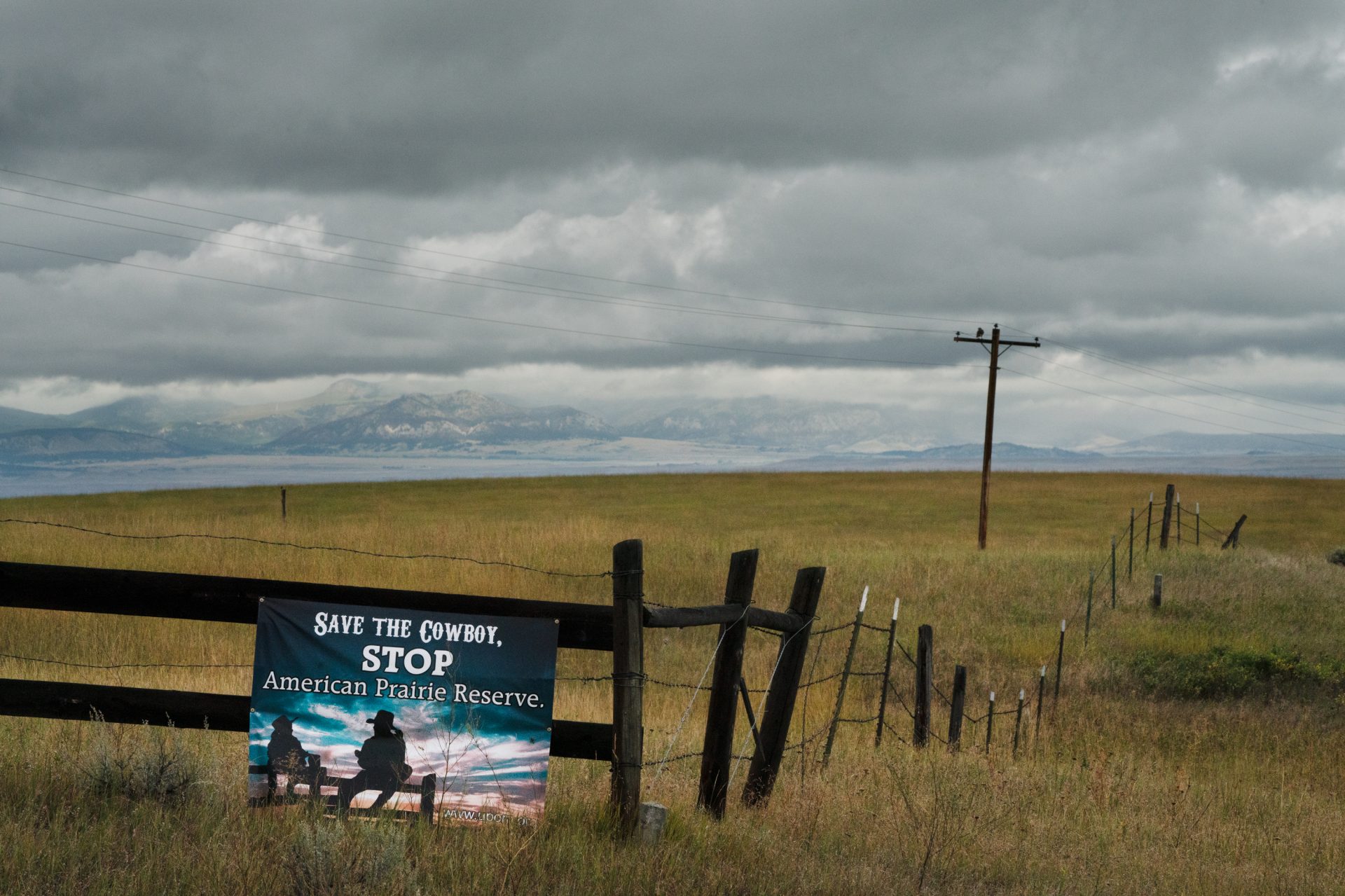 A "Save the Cowboy" sign is posted along a fence. The "Little Rockies" on the Fort Belknap Indian Reservation are seen in the distance.