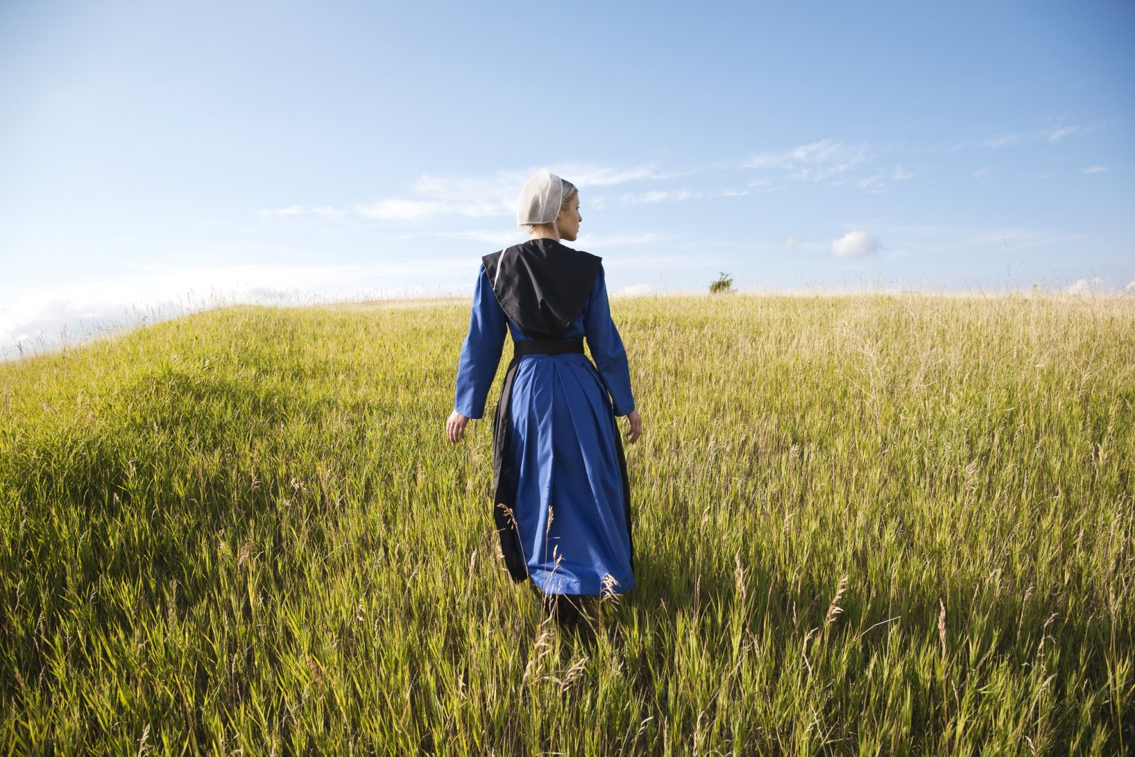An Old Order Amish woman in a blue dress and black cape and apron walks in a grassy field on a sunny afternoon