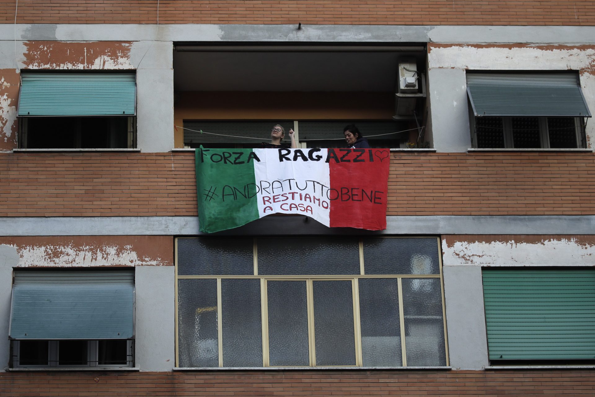 People stand on a balcony adorned with an Italian flag during one of the many flash mobs taking place these days in Rome, Sunday, March 15, 2020. The nationwide lockdown to slow coronavirus is still early days for much of Italy, but Italians are already showing signs of solidarity with flash mob calls circulating on social media for people to ''gather'' on their balconies at certain hours, either to play music or to give each other a round of applause. For most people, the new coronavirus causes only mild or moderate symptoms. For some, it can cause more severe illness, especially in older adults and people with existing health problems.