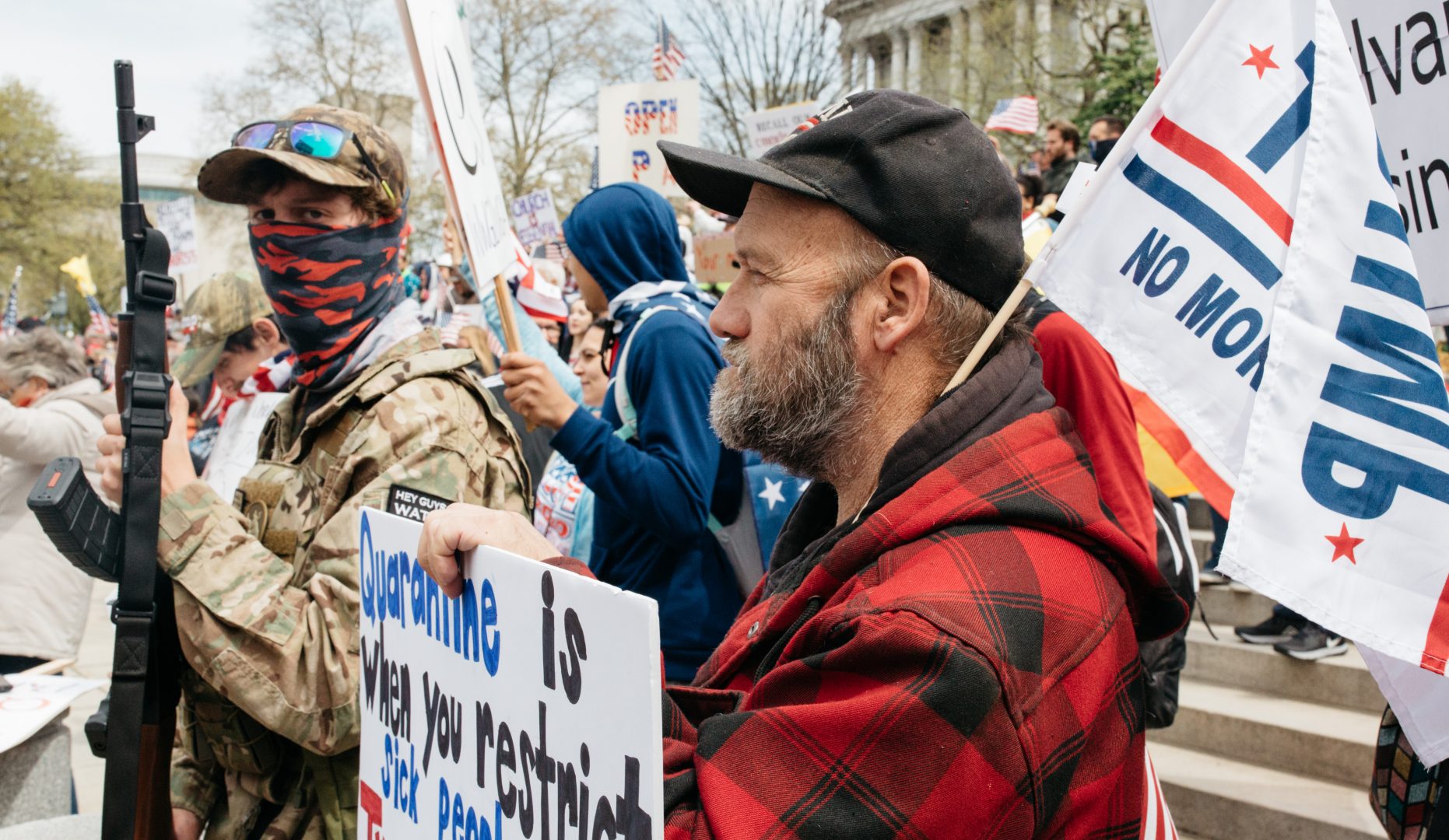 At right, a man with a Trump flag holds a sign protesting the state shutdown. At left, a masked man holds an AK-47-style rifle. 