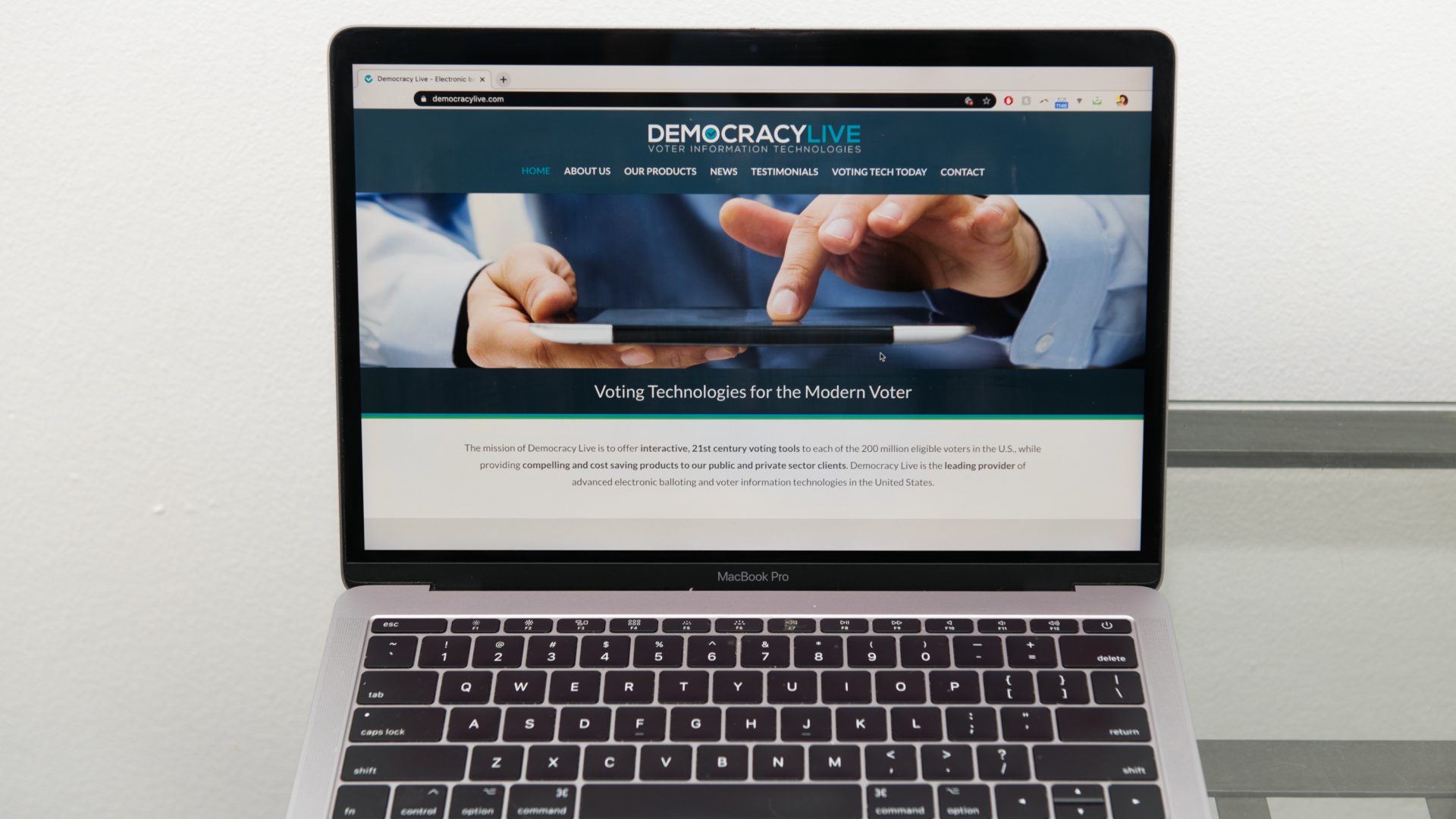 The Democracy Live home page is displayed on an Apple laptop computer. The company is administering a ballot return system for disabled voters in West Virginia, Delaware, and potentially New Jersey.