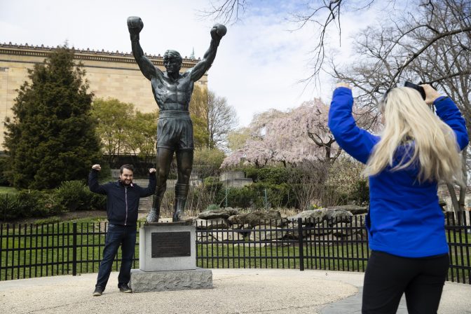 In this April 3, 2020, photo, Jessica Shiroff, right, directs exchange student Joao Martucci, of Brazil, as he poses for a photograph with the Rocky Statue at the Philadelphia Art Museum in Philadelphia. “Rocky” finished tied for No. 2 in The Associated Press Top 25 favorite sports movies poll.