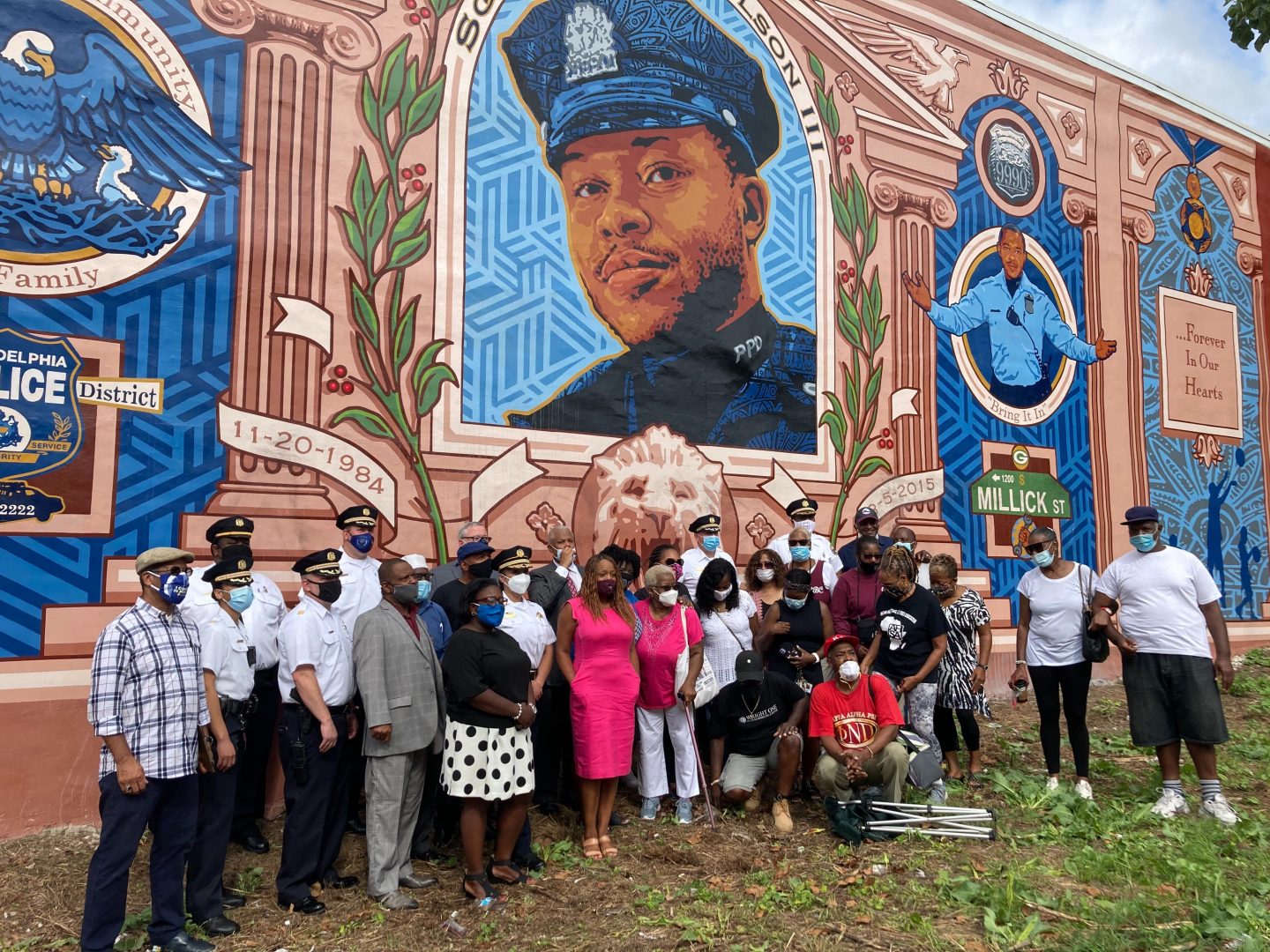 City officials, police officers from the 18th district, and neighborhood residents pose in front of the mural on 61st Street and Baltimore Avenue, dedicated to Sgt. Robert Wilson III.