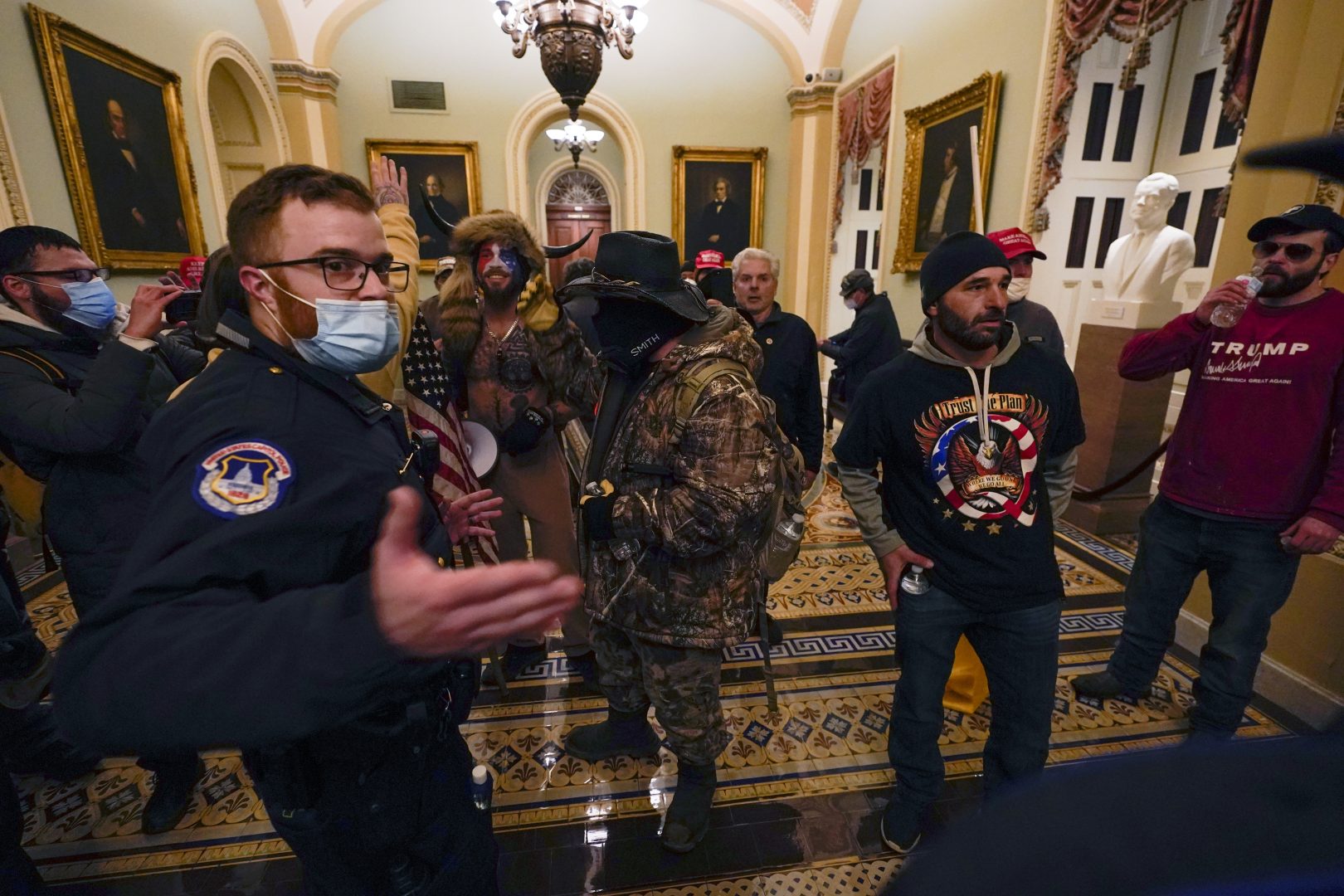 Rioters walk as U.S. Capitol Police officers watch in a hallway near the Senate chamber at the Capitol in Washington, Wednesday, Jan. 6, 2021. A court filing against Jacob Chansley, seen here wearing face paint and fur, states the mob wanted to 