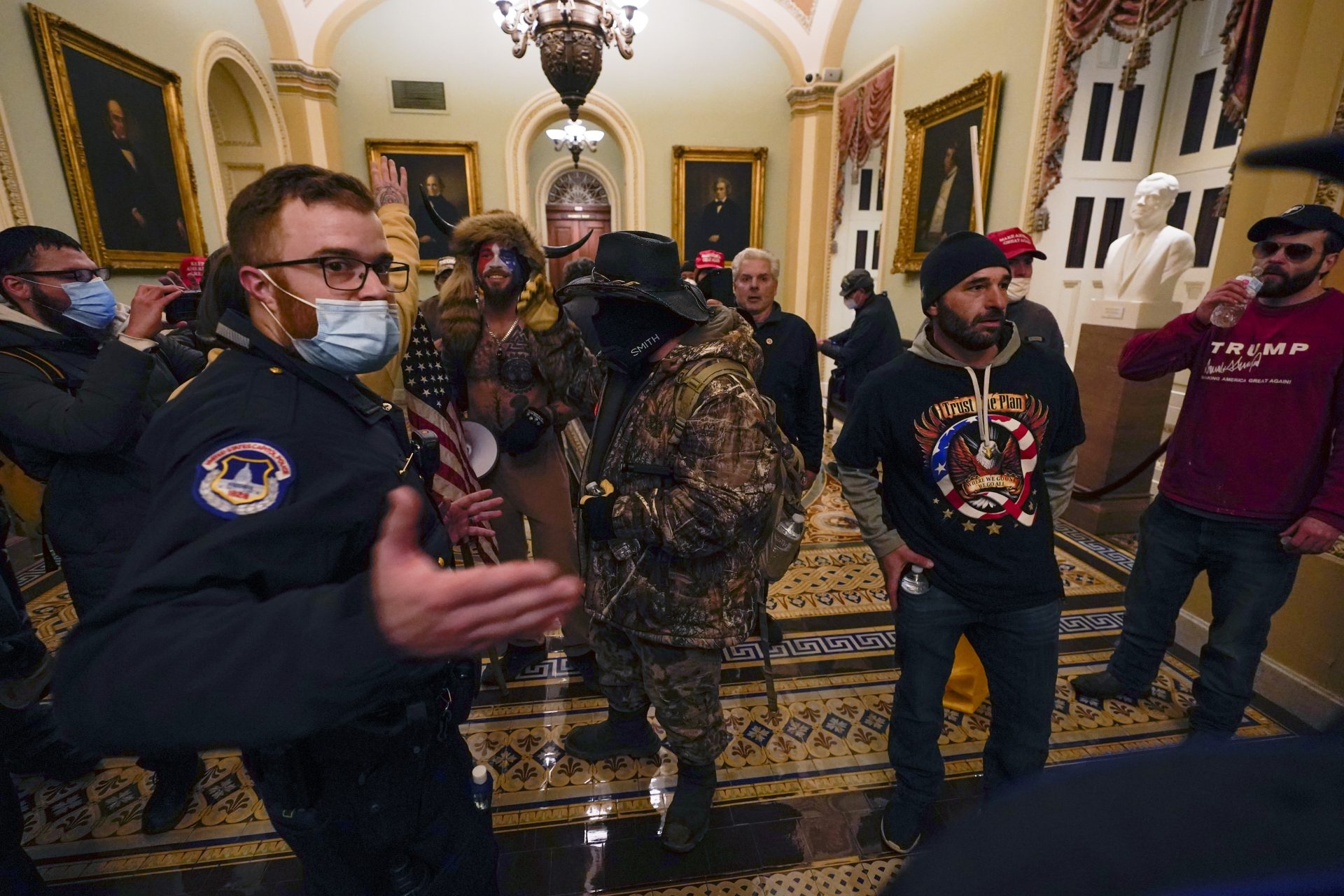 Rioters walk as U.S. Capitol Police officers watch in a hallway near the Senate chamber at the Capitol in Washington, Wednesday, Jan. 6, 2021. A court filing against Jacob Chansley, seen here wearing face paint and fur, states the mob wanted to "capture and assassinate elected officials in the United States Government."