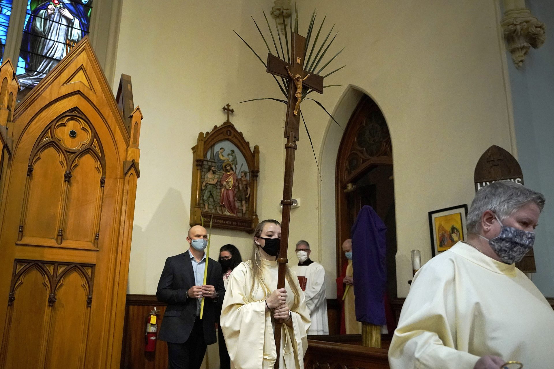 In this Sunday, March 28, 2021 file photo, altar server Samantha Holmes, of East Brookfield, Mass., center, carries a crucifix with palm leaves during a procession at the start of Palm Sunday Mass at Mary, Queen of the Rosary Parish in Spencer, Mass.
