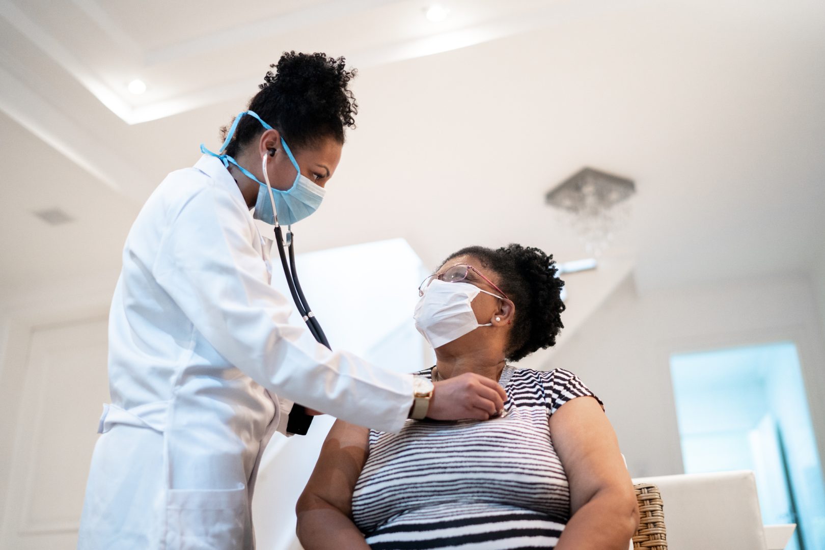 Doctor listening to patient's heartbeat during home visit - wearing face mask