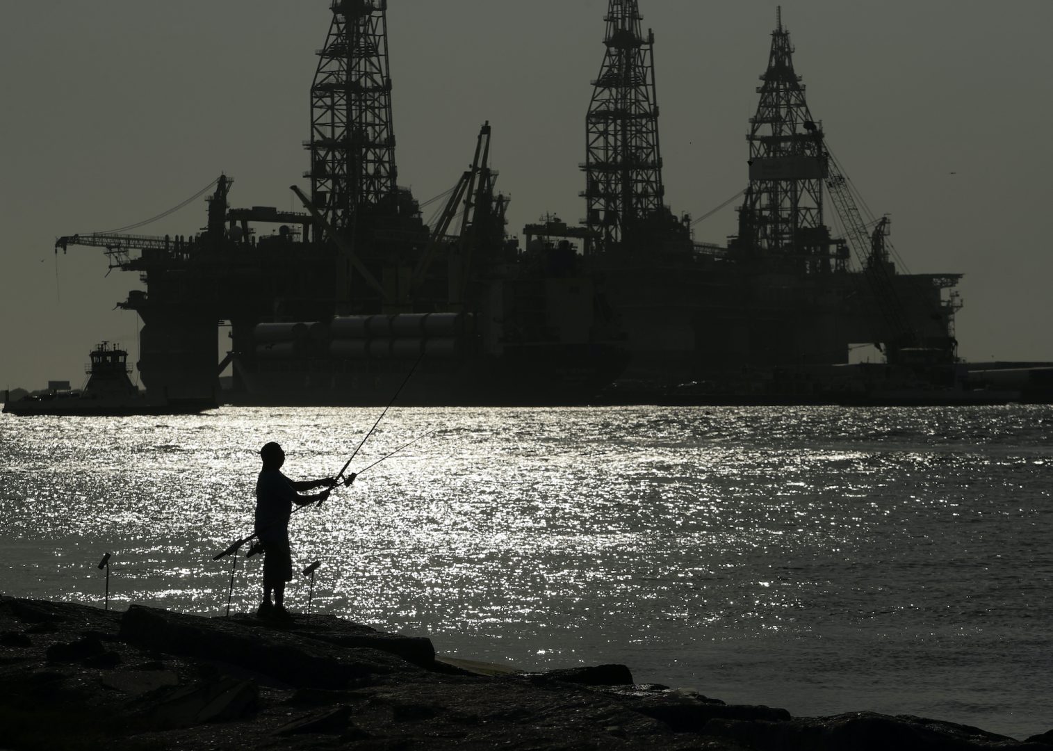 FILE - A man wears a face mark as he fishes near docked oil drilling platforms, Friday, May 8, 2020, in Port Aransas, Texas. The U.S. Interior Department on Wednesday, Nov. 17, 2021, is auctioning vast oil reserves in the Gulf of Mexico estimated to hold up to 1.1 billion barrels of crude. It's the first such sale under President Joe Biden and underscores the challenges he faces to reach climate goals that rely on cuts in fossil fuel emissions.  