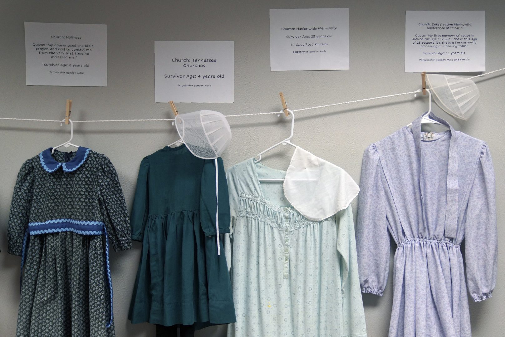 Dresses donated by sexual assault survivors from Amish and other plain-dressing religious groups hang on a clothesline beneath a description of each survivors' age and church affiliation, on Friday, April 29, 2022, in Leola, Pa. The exhibit's purpose was to show that sexual assault is a reality among children and adults in such groups. Similar exhibits held nationwide aim to shatter the myth that abuse is caused by a victim's clothing choice. (AP Photo/Jessie Wardarski)