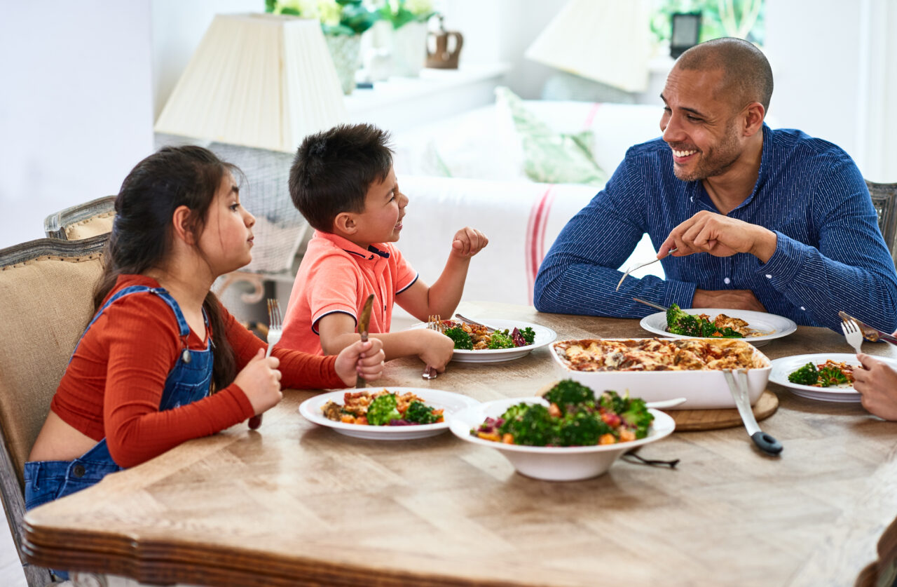 Mature man listening to son and daughter, enjoying healthy home cooked dinner, fresh food in dishes on dining room table
