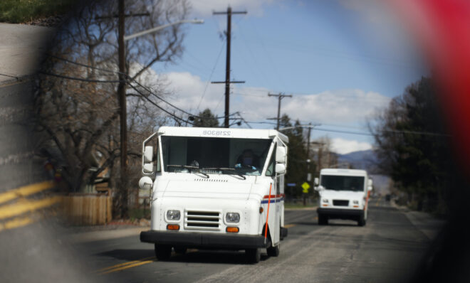 United States Post Office delivery trucks are reflected in the side mirror of a vehicle as postal delivers set off on their daily rounds  as a statewide stay-at-home order remains in effect in an effort to reduce the spread of the new coronavirus Tuesday, March 31, 2020, in  Arvada, Colo. The new coronavirus causes mild or moderate symptoms for most people, but for some, especially older adults and people with existing health problems, it can cause more severe illness or death. (AP Photo/David Zalubowski)