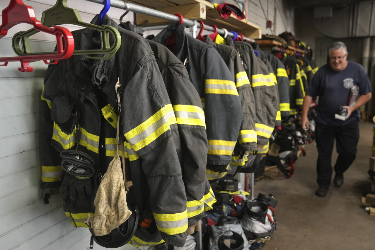 A Brockton firefighter walks past protective gear at Station 1, Thursday, Aug. 3, 2023, in Brockton, Mass. Firefighters around the country are concerned that gear laced with the toxic industrial compound PFAS could be one reason why cancer rates among their ranks are rising. The chemical, which has been linked to health problems including several types of cancer, is used in turnout gear to repel water and other substances when fighting a fire. (AP Photo/Steven Senne)