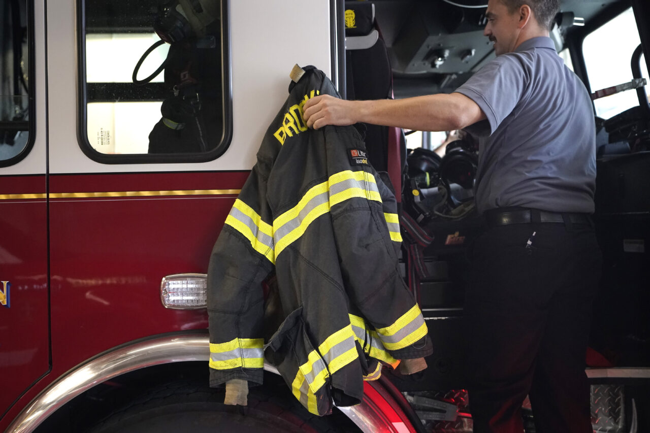A Brockton firefighter lifts a protective turnout coat onto a firetruck at Station 1, Thursday, Aug. 3, 2023, in Brockton, Mass. Firefighters around the country are concerned that gear laced with the toxic industrial compound PFAS could be one reason why cancer rates among their ranks are rising. The chemical, which has been linked to health problems including several types of cancer, is used in turnout gear to repel water and other substances when fighting a fire. (AP Photo/Steven Senne)