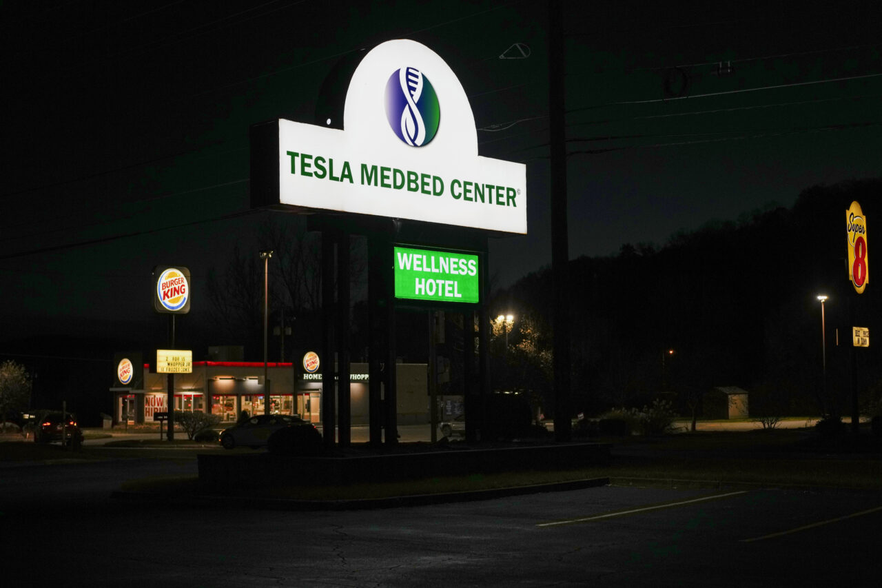 An illuminated sign across from Burger King and a Super 8 Hotel shows the entrance for Tesla BioHealing's Tesla Wellness Hotel and MedBed Center on Tuesday, Nov. 14, 2023, in Butler, Pa. Part motel, part new-age clinic, the facility offers nightly rentals in rooms that come equipped with “biohealers” – small canisters that the company claims exude “life force energy,” or biophotons. (AP Photo/Carolyn Kaster)