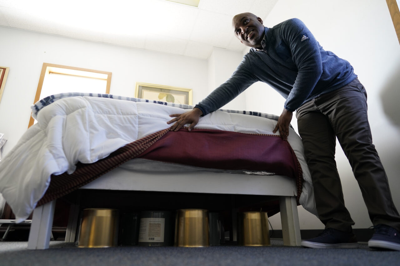 Seth Robinson, a chiropractor who serves as Tesla BioHealing's medical director, lifts bed skirts to show "biohealers" – canisters that the company claims exude "life force energy," or biophotons, at the Tesla BioHealing & MedBed Center, in Milford, Del., Tuesday, Oct. 3, 2023. According to QAnon adherents, medbeds were developed by the military (in some versions, using alien technology) and are already in use by the world’s richest and most powerful families. (AP Photo/Carolyn Kaster)