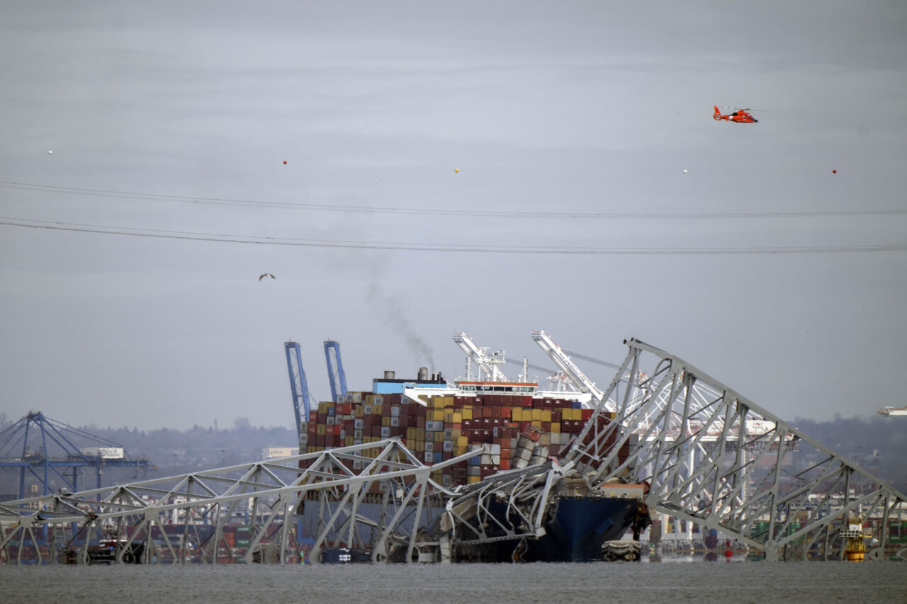 A helicopter flies over a container ship as it rests against wreckage of the Francis Scott Key Bridge on Tuesday, March 26, 2024, as seen from Pasadena, Md. The container ship lost power and rammed into the major bridge in Baltimore early Tuesday, causing it to snap and plunge into the river below. Several vehicles fell into the chilly waters, and rescuers searched for survivors. (AP Photo/Mark Schiefelbein)